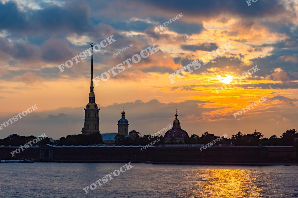 На фотографии изображена Петропавловская крепость на фоне живописного заката в Санкт-Петербурге. Шпиль Петропавловского собора возвышается над крепостью, добавляя величественности этому историческому месту. Закатное небо, окрашенное в тёплые тона, создает спокойную и романтичную атмосферу, а вода реки Невы отражает цвета неба, усиливая эффект. Этот вид — одна из знаковых сцен вечернего Петербурга.